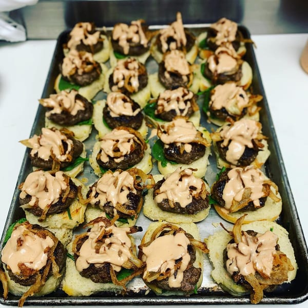 A tray of mini burgers with fried onions, spinach, and a creamy sauce on buns.